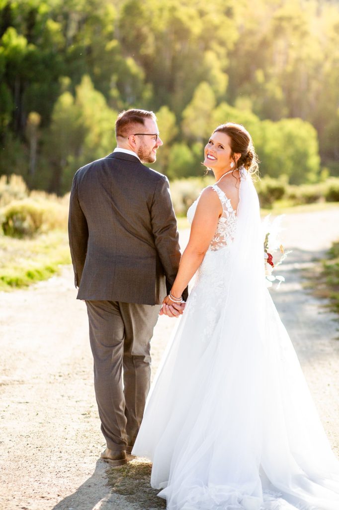 Bride in a white wedding gown and veil holding hands with a groom in a gray suit walking away on a sunlit outdoor path Colorado Wedding Photographer Colorado Wedding Photographer