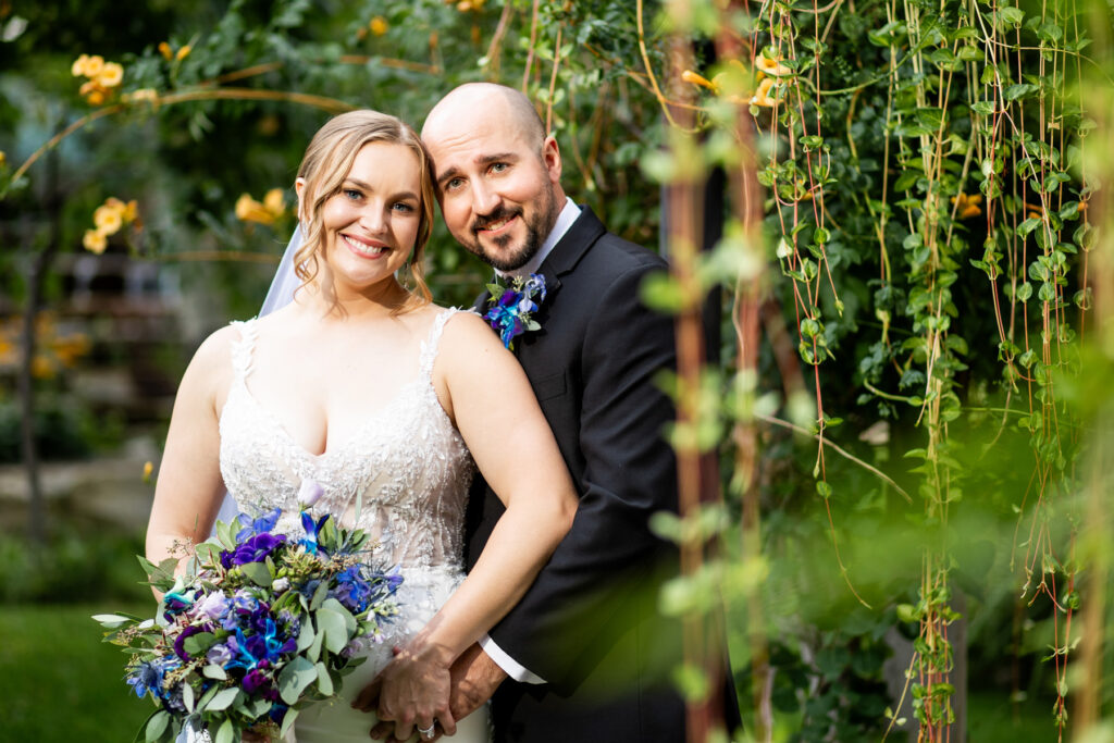 Bride and groom posing together in a garden the bride in a lace white dress holding a bluepurple bouquet both smiling warmly Colorado Wedding Photographer Colorado Wedding Photographer