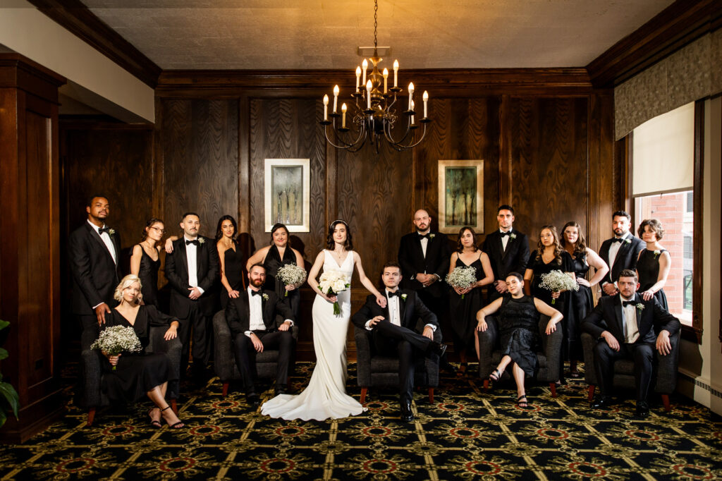 Bride in a white gown stands center with bridesmaids and groomsmen in black posing in a wood paneled room with a chandelier above the group Colorado Wedding Photographer Colorado Wedding Photographer