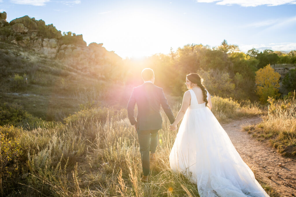 Bride in a white wedding dress and groom in a suit walk hand in hand along a sunlit dirt path at golden hour field and rocks in the background Colorado Wedding Photographer