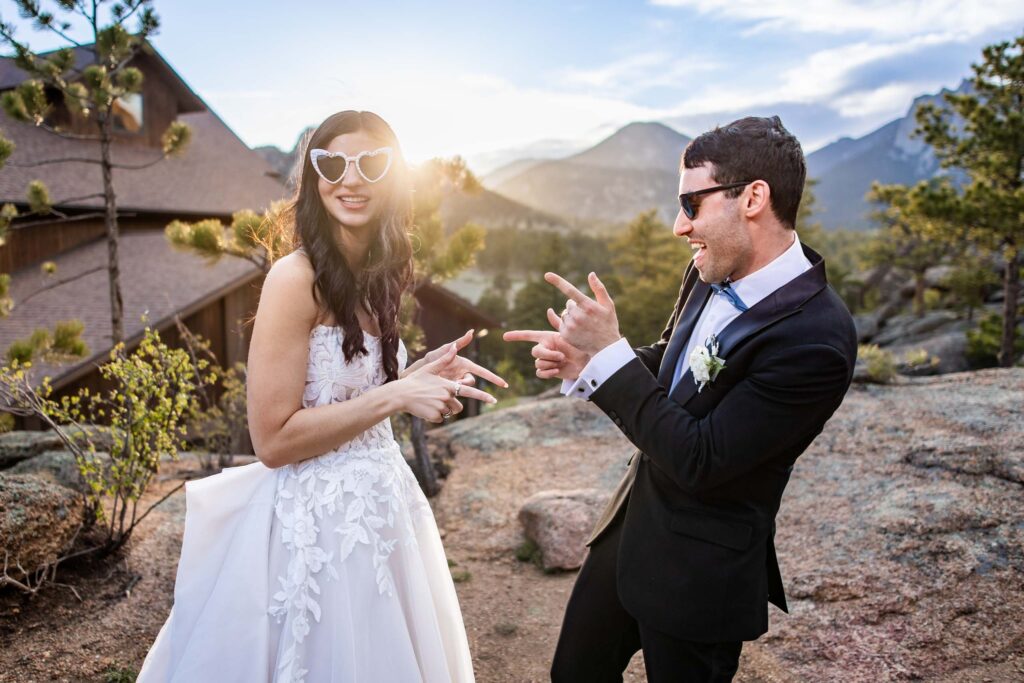 Bride in a lace wedding dress and groom in a dark suit playfully pointing at each other outdoors with mountains in the background sun setting Colorado Wedding Photographer Colorado Wedding Photographer