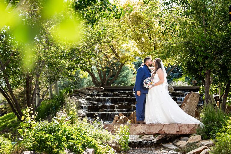 Bride in a white wedding gown and a groom in a blue suit share a kiss on a stone bridge surrounded by lush greenery and a small waterfall in a sunlit garden Colorado Wedding Photographer Colorado Wedding Photographer