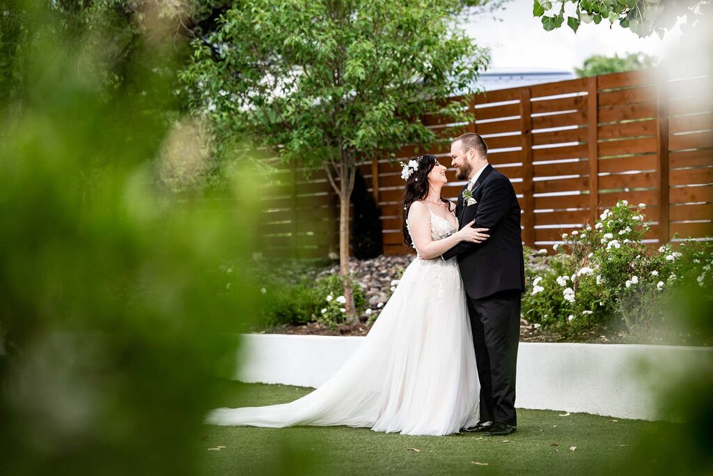 Bride in a white wedding gown and groom in a black suit embrace in a garden with a wooden fence and white flowers behind them Colorado Wedding Photographer Colorado Wedding Photographer