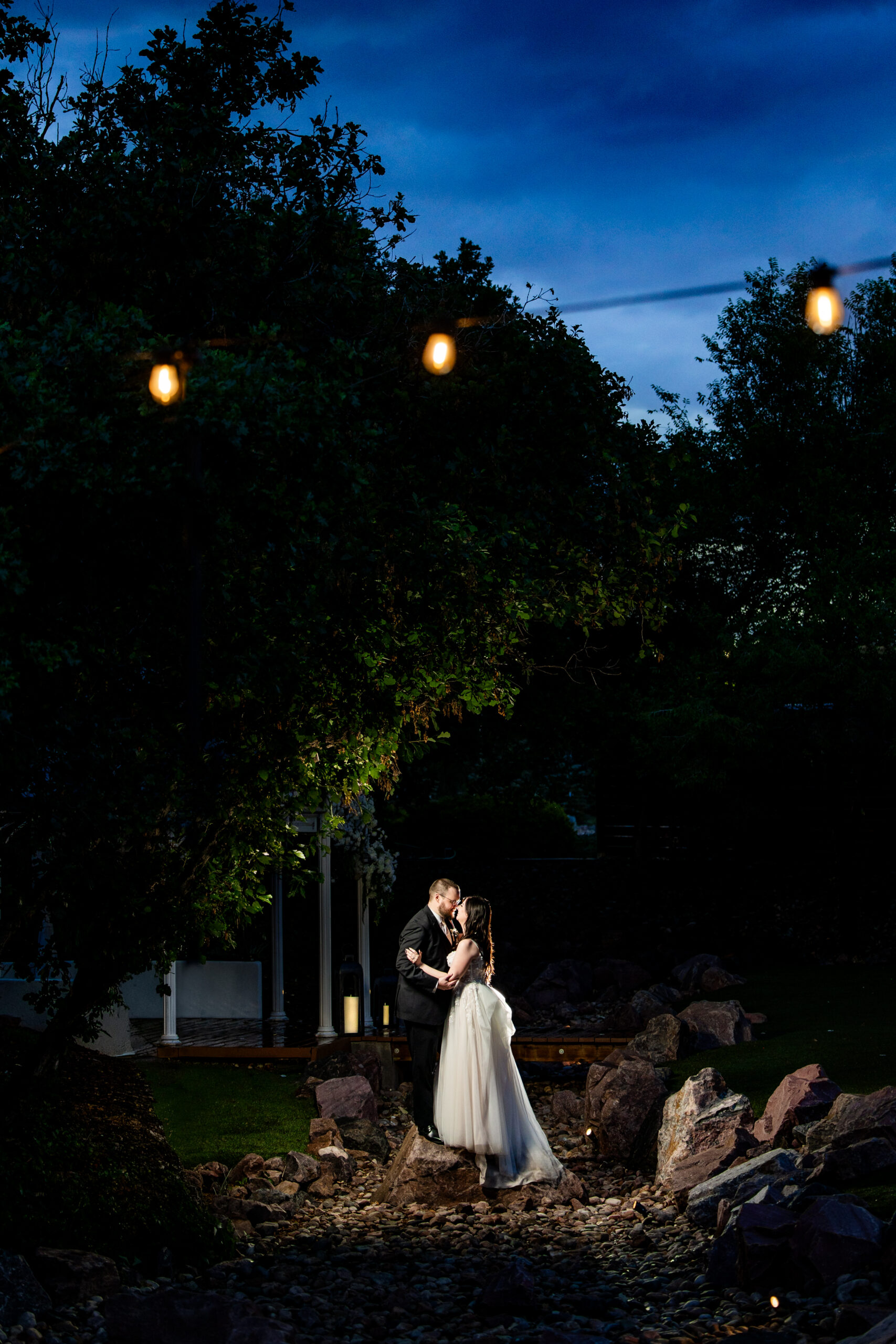 Bride and groom share a kiss in a rocky garden at dusk warm string lights glowing above them Colorado Wedding Photographer Colorado Wedding Photographer