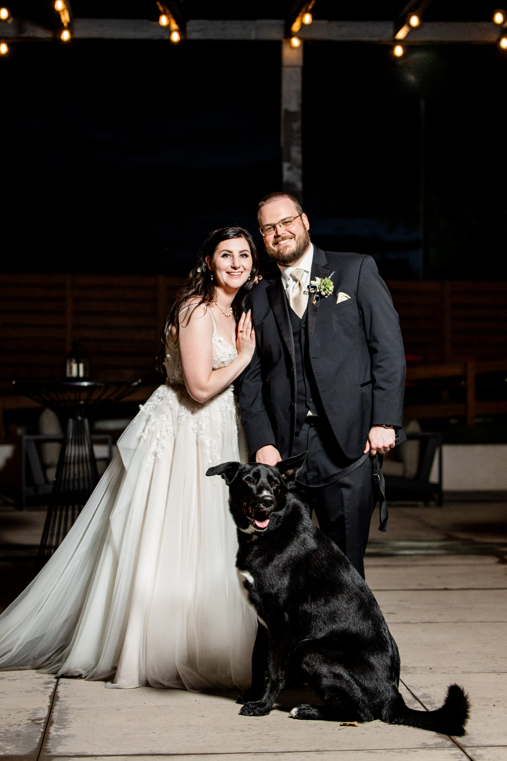 Bride and groom in formal wear pose at night with a large black dog seated in front of them under string lights Colorado Wedding Photographer Colorado Wedding Photographer