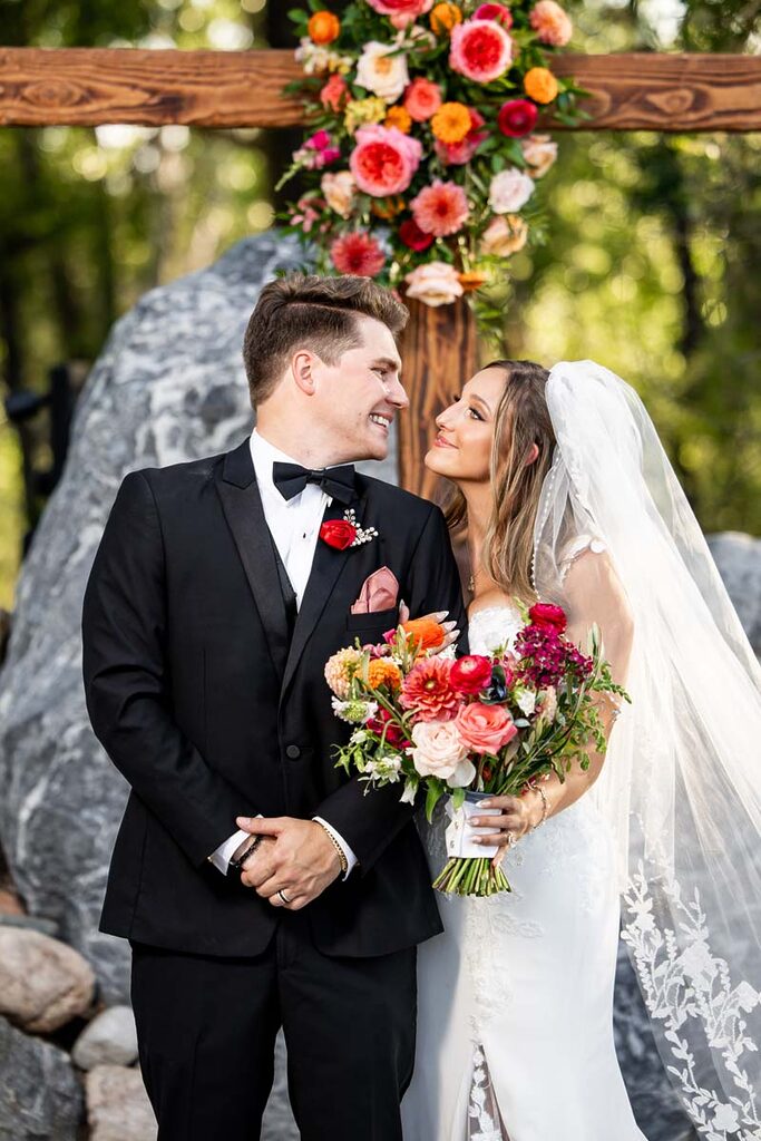 Bride and groom in formal wear smile at each other under a colorful flower arch bouquet in the foreground Colorado Wedding Photographer Colorado Wedding Photographer