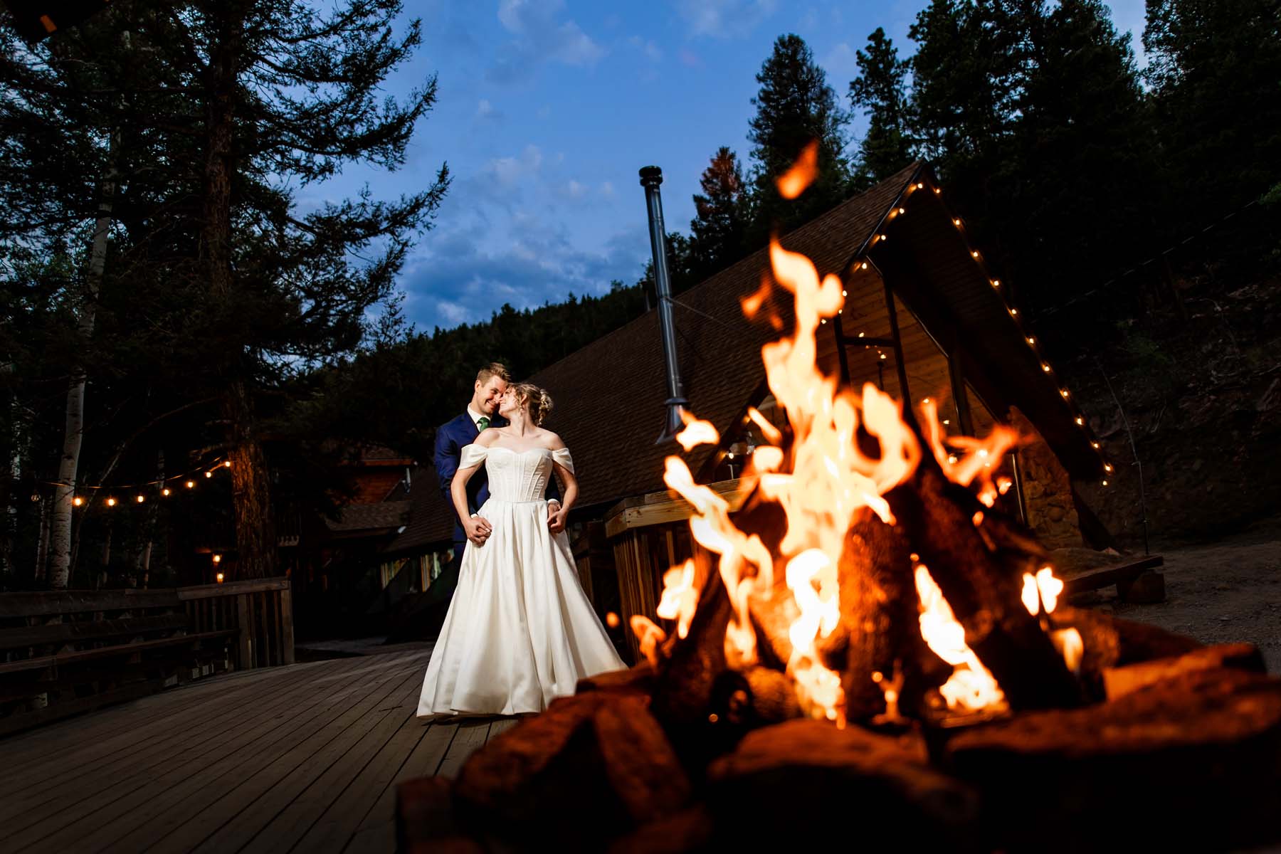 A bride and groom in white wedding dress and dark suit stand close by a large bonfire outdoors at dusk with string lights on a wooden cabin in the background Colorado Wedding Photographer Colorado Wedding Photographer