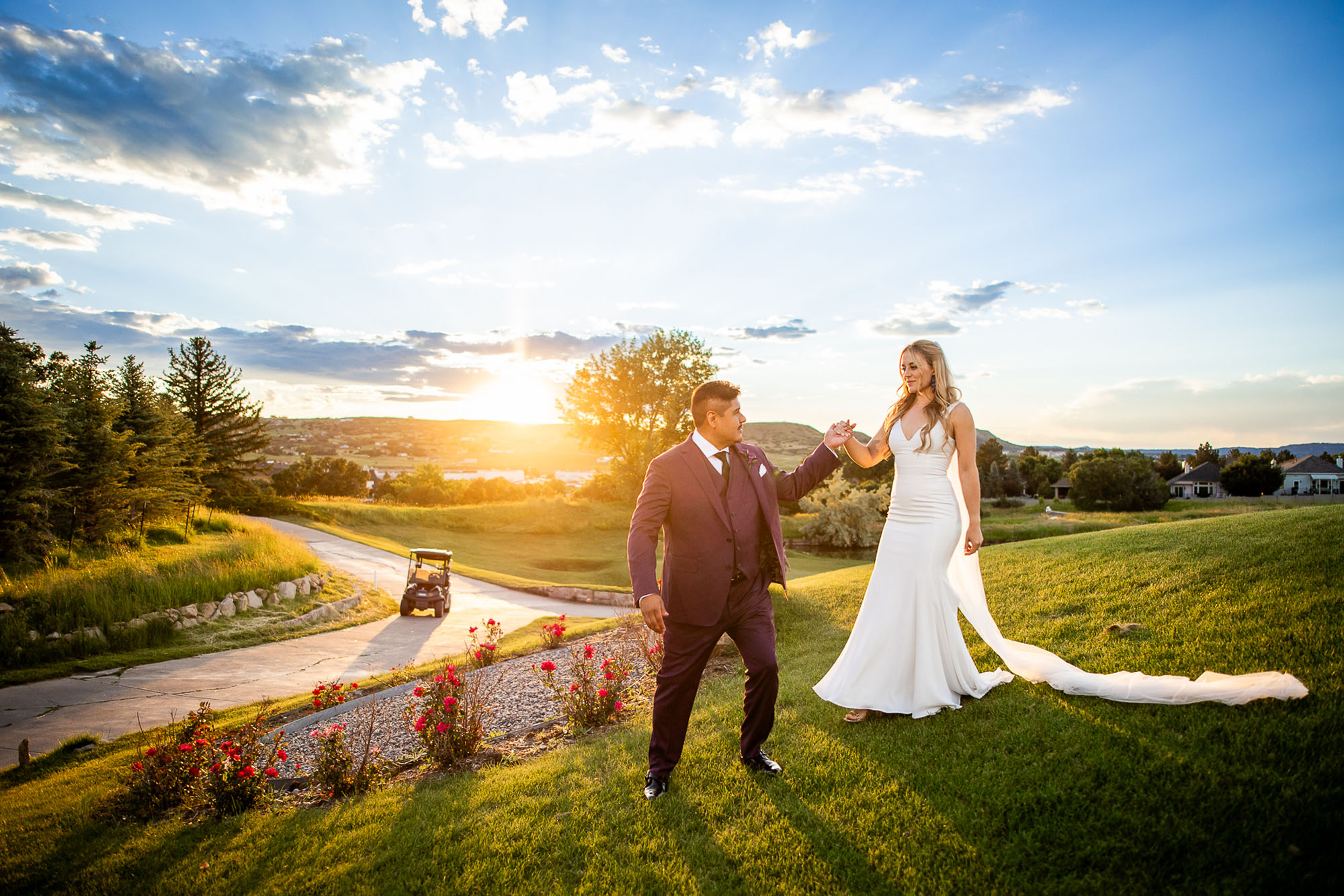 Sarah-Goff-Photography-Castle-Rock-Summer-Wedding-Oaks-at-Plum-Creek-Golf-Venue 101 - Colorado Wedding Photographer Bride in a white wedding gown and groom in a burgundy suit walk hand in hand on a sunlit lawn at sunset with a golf cart in the background Colorado Wedding Photographer