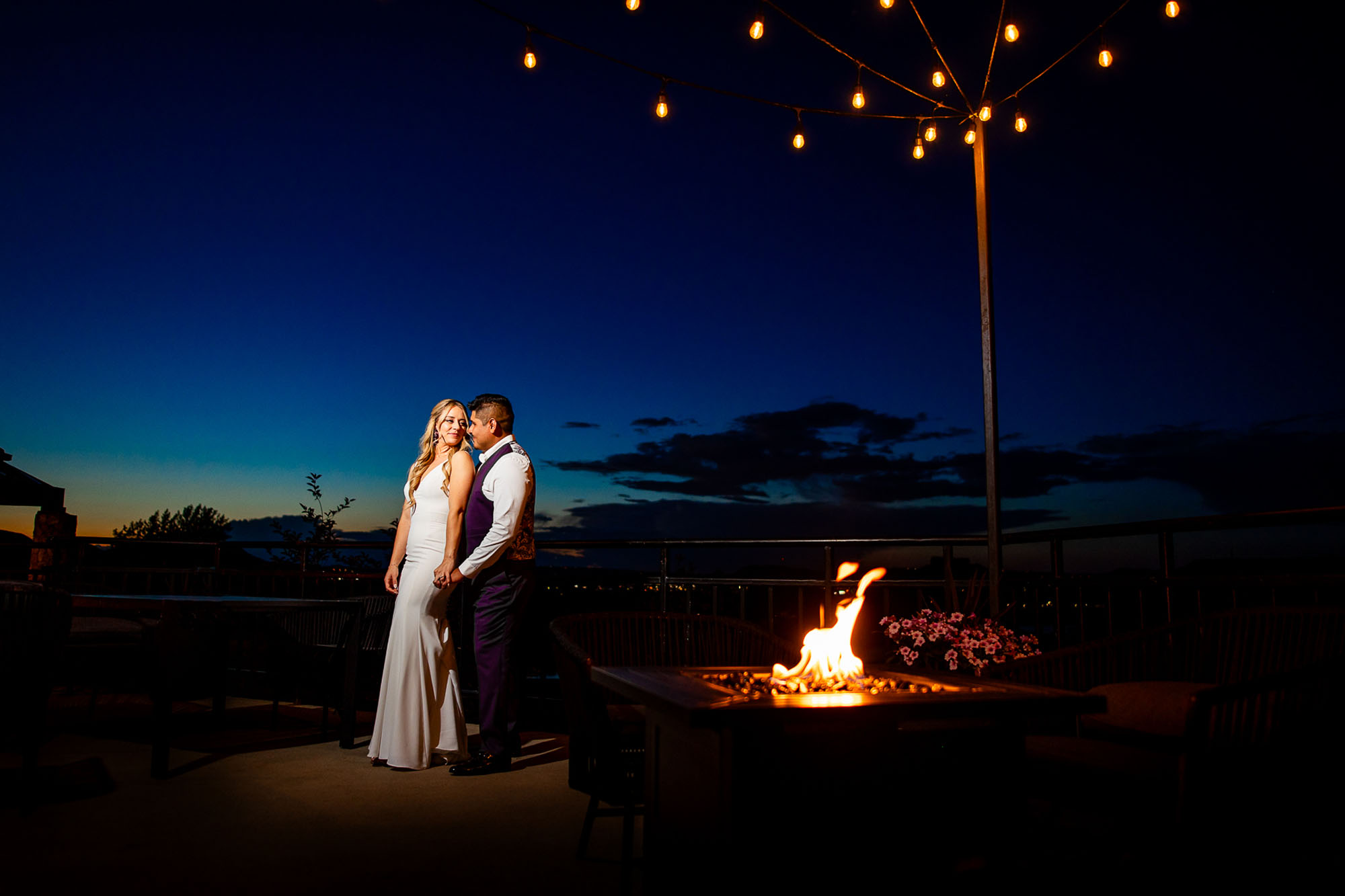 A couple in formal attire stands close together by a glowing fire pit on an outdoor deck at dusk with warm string lights overhead Colorado Wedding Photographer Colorado Wedding Photographer